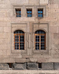 Stone facade with barred rectangular and arched windows, featuring carved frames and stylized geometric patterns.