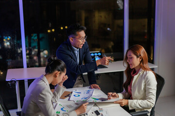 Business team members working overtime in a modern office, analyzing financial charts and discussing strategy on a late night