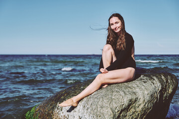 Young woman sitting on rock by Baltic Sea under clear blue sky
