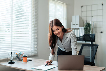 Young Asian businesswoman working at her desk. She is reviewing documents while checking data on a laptop in a modern office