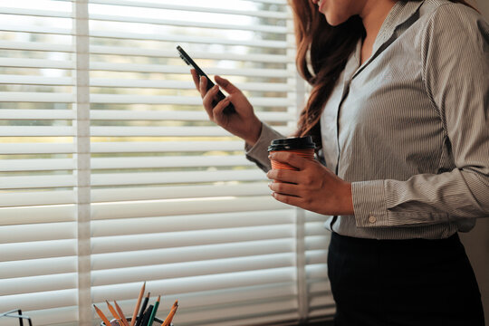 Businesswoman taking a coffee break, holding a disposable cup and checking her smartphone near an office window with blinds - Powered by Adobe