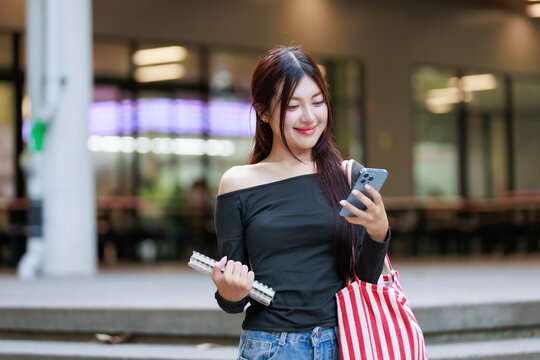 Smiling young adult asian woman student holding notebook and tote bag, checking smartphone on university campus - Powered by Adobe