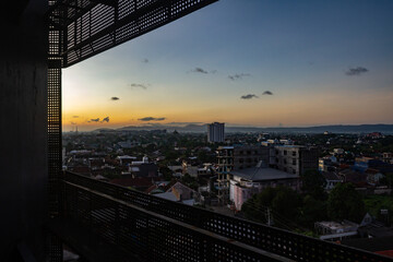 A high-angle view of a dense urban landscape at twilight, framed by a silhouetted perforated metal structure in the foreground. The skyline features residential rooftops, mid-rise buildings, and dista