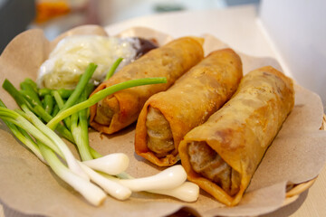 Three deep-fried Lumpia Semarang spring rolls rest on greaseproof paper, accompanied by fresh Allium fistulosum stalks and dipping sauces. The golden pastry wrappers encase a savory bamboo shoot filli