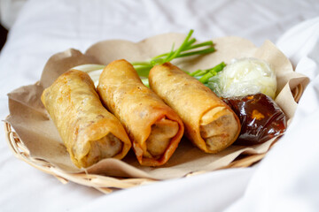 Three deep-fried Lumpia Semarang spring rolls sit on brown paper in a woven basket, served with fresh Allium fistulosum, pickled cucumber in plastic, and thick dipping sauce on a white surface.