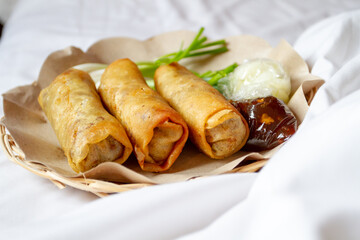 Three deep-fried Lumpia Semarang spring rolls sit on brown paper in a woven basket, served with fresh Allium fistulosum, pickled cucumber in plastic, and thick dipping sauce on a white surface.