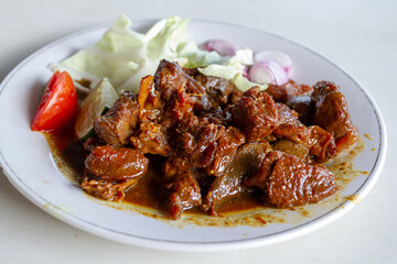 A plate of Indonesian fried goat meat, widely known as Sate Goreng, served in sweet soy sauce. The savory dish is garnished with fresh cabbage, sliced red shallots, tomato, and lime on a white plate.