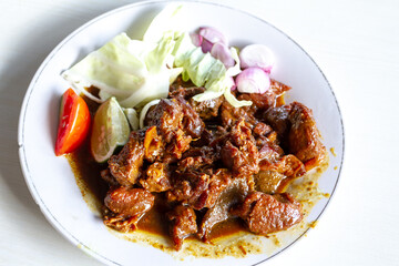 A plate of Indonesian fried goat meat, widely known as Sate Goreng, served in sweet soy sauce. The savory dish is garnished with fresh cabbage, sliced red shallots, tomato, and lime on a white plate.