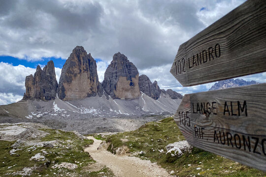 Wooden wayfinding signs for Landro, Lange Alm and Rifugio Auronzo on the Tre Cime loop; rocky path toward the three towers under a partly cloudy sky in Italy