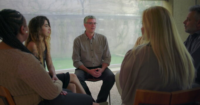 Middle aged man sharing emotional testimony during therapy group session, participants listening attentively in circle during vulnerable moment of support and healing