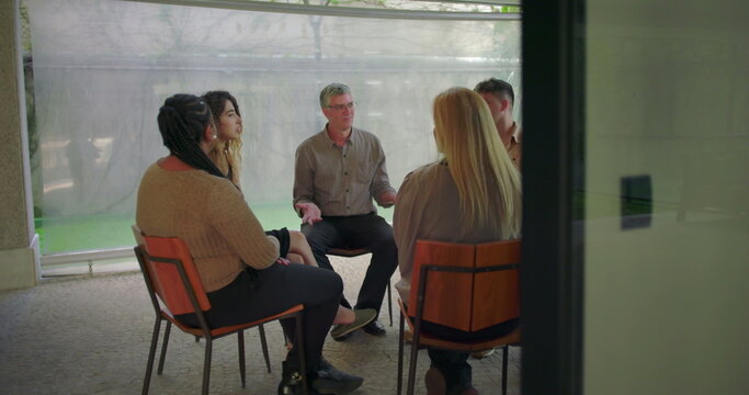 Middle aged man sharing emotional testimony during therapy group session, participants listening attentively in circle during vulnerable moment of support and healing