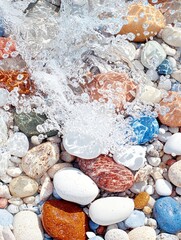 Close-up of water splashing over colorful pebbles on a beach. The water is clear, and the pebbles are smooth and varied in color. The scene is bright and sunny.