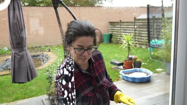 Woman with gloves and glasses cleans glass door with squeegee in back yard on a bright day using soapy water