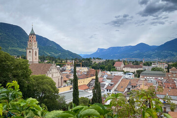 Cityscape of Merano in South Tyrol, Italy with St. Nicholas Church and bell tower above rooftops; green hills and distant mountains under a sky with clouds