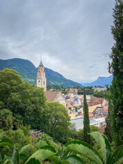 Cityscape of Merano in South Tyrol, Italy with St. Nicholas Church and bell tower above rooftops; green hills and distant mountains under a sky with clouds