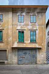 Weathered historic building facade with peeling plaster, tall windows and a heavy metal door; muted tones and urban texture on a quiet street in Italy