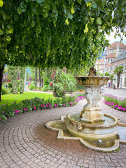 Ornate stone fountain with flowerbeds and palms on the Passer Promenade in Merano (Meran), Italy; landscaped paths near the Kurhaus under a sky with cloud