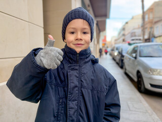 Cute little smiling boy wearing winter clothes showing thumb up in a winter day in the city. School boy.