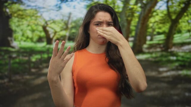 Young hispanic woman wearing an orange sleeveless top in green park pinches her nose to block a strong outdoor odor; disgust.