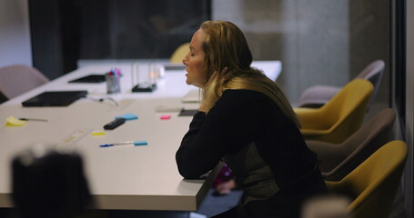 Middle-aged businesswoman leans forward with head in hand at cluttered conference table, exhausted...