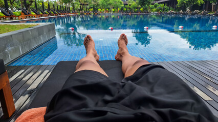 Relaxing poolside view with a person lying on a black sun lounger, overlooking a large blue-tiled swimming pool surrounded by greenery and resort buildings.