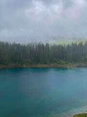 Rain softens the turquoise water of Lake Carezza (germ. Karersee) beneath spruce and fir forest in South Tyrol, Italy, with low mist drifting across the Dolomites