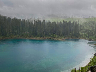 Rain softens the turquoise water of Lake Carezza (germ. Karersee) beneath spruce and fir forest in South Tyrol, Italy, with low mist drifting across the Dolomites