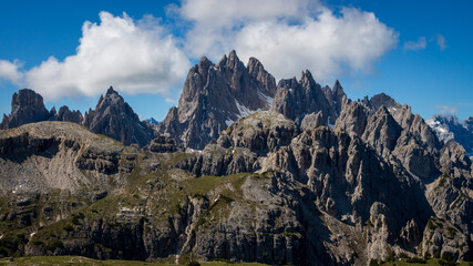 Scenic view of Cadini di Misurina mountain group seen from the Tre Cime area in Italy;  jagged Dolomite spires, layered cliffs and green slopes with small snow patches against blue sky with clouds
