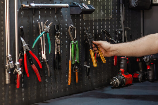 Mechanic's hand reaching for wire strippers on a pegboard wall. Organized workshop with wrenches, pliers, and impact driver.