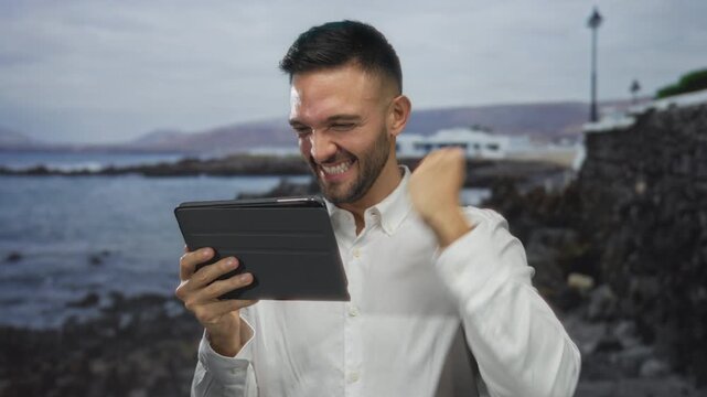Young man with tablet smiling on a beach with a scenic seashore in the background showcasing technology and travel outdoors in a vibrant coastal setting.