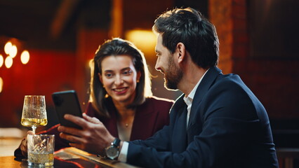Flirting lady talking man at night inside pub lounge. Couple looking smartphone