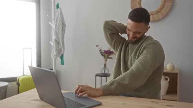 Young man with laptop indoors experiencing neck pain in a cozy living room with green sweater and modern decor including a wooden table and a decorative mirror on the wall.