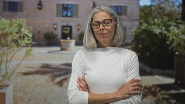 Woman with gray hair and glasses, arms crossed, standing in front of stone building courtyard near a fountain outdoors with slight smile; maturity confidence.