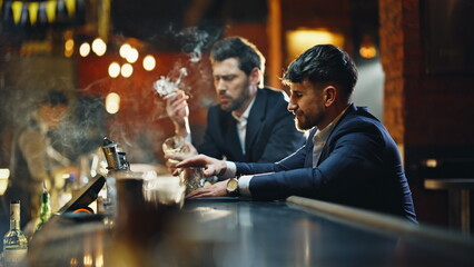 Two men drinking bar puffing smoke sitting in amber illuminated light closeup