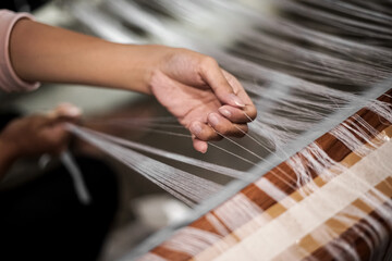 A hand gently pulling weaving threads on a loom as part of fabric preparation in a textile workspace.