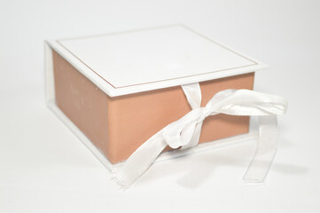 Studio shot of a small, square gift box with a white lid and salmon-colored base, tied neatly with a white satin ribbon bow, isolated on a clean white background. Concept for holiday, luxury packaging