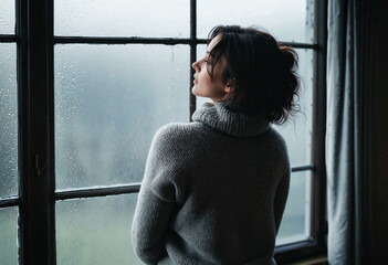 A woman stands by a window, gazing outside. She wears a gray turtleneck sweater and has her hair loosely tied up. The window is covered in raindrops, creating a misty background. 