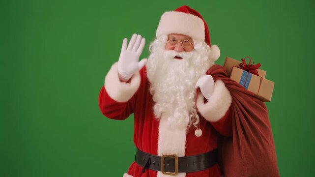 Santa Claus in a red suit with a white beard waving and holding a bag of gifts against a green background.