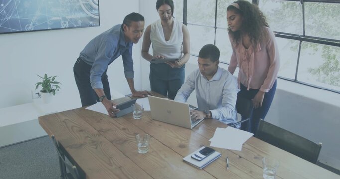 Collaborating team in shirts and blouses leaning over silver laptop in meeting room, viewing tablet