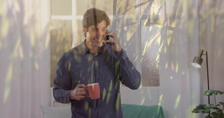 Holding orange mug, man wearing denim shirt smiling, speaking on smartphone in living room, plant