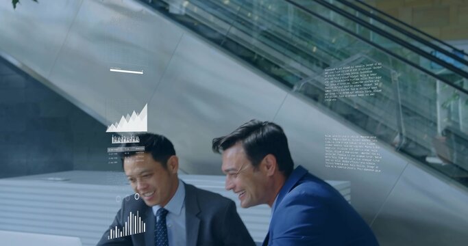 Leaning two men in business suits examining laptop at corporate atrium, with holographic charts
