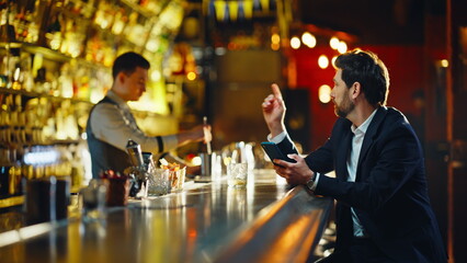 Rich businessman scrolling mobile screen at bar counter. Guest sipping brandy