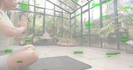 Kneeling woman pressing palms on dark yoga mat in sunroom, in top, shorts, plants, copy space