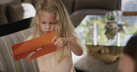 Holding orange craft paper, blonde child studying cut strips at home, wearing peach sleeveless top