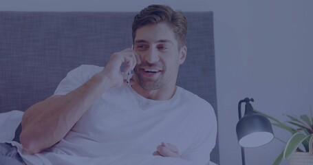 Smiling mid-adult man reclining on bed in bedroom, holding smartphone and wearing white T-shirt