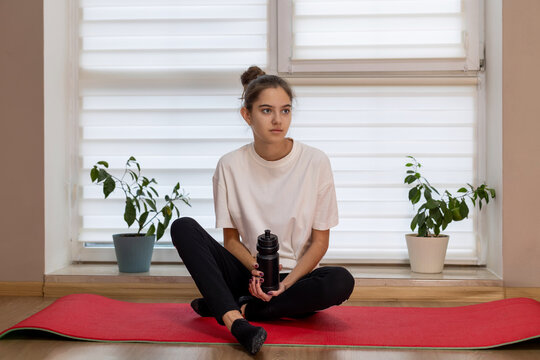 Teen girl resting after home workout session. Young girl sitting on a yoga mat with a water bottle, taking a break from exercising in a bright indoor home setting.