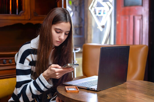 Teen girl focused on smartphone at cafe table. Young girl sitting at a wooden table in a cozy cafe, concentrating on her phone with an open laptop in front of her.