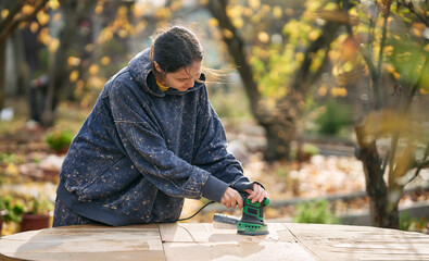 Young woman sanding wooden table in autumn garden