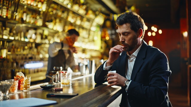 Weary man lighting cigarette sitting bar with half empty glass nearby closeup