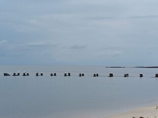 The wooden beams of an old wooden jetty protruding from the tranquil bay water on a cloudy day at the beach.